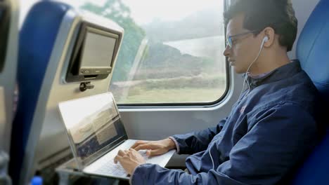 a young software engineer wearing eyeglasses working on a laptop and listing to music in moving train