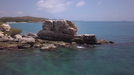 aerial: a tracking shot around a high stone formation and rocks sumberged in the azure ocean, an island and mountain range is visible as the drone rotates around the rock