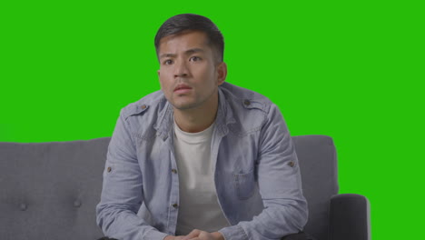 studio shot of tense young man sitting on sofa watching sport on tv against green screen