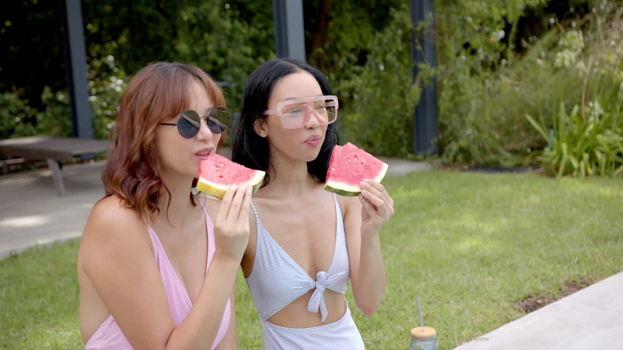 Two young biracial female friends enjoy watermelon by pool, celebrating friendship