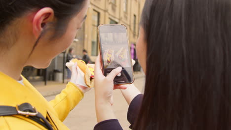 dos amigas tomando fotos de perritos calientes comprados en un puesto de comida callejera en un teléfono móvil
