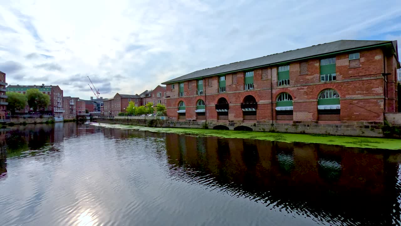 Smooth daytime pan along a river in Leeds, showing historic brick buildings, modern apartments, and reflections in calm water under cloudy skies