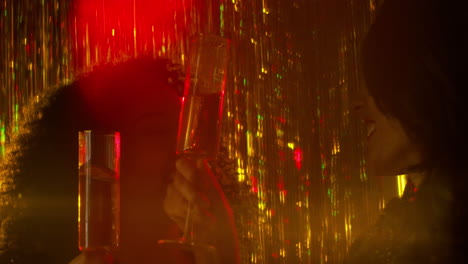 close up of two women in nightclub or bar celebrating doing cheers and drinking alcohol with sparkling lights