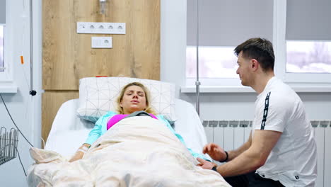 blonde pregnant lady lies in bed and breathes deep to relief pain from contractions. man sitting beside strokes her to sooth.