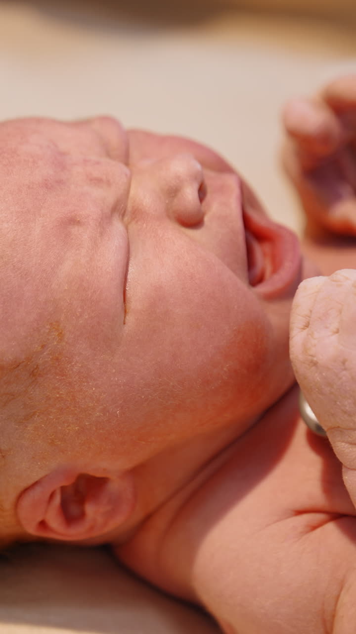Newborn red-haired baby cries while doctor's gloved hand puts stethoscope to its chest. Checking the life of a child after birth. Close up. Vertical video
