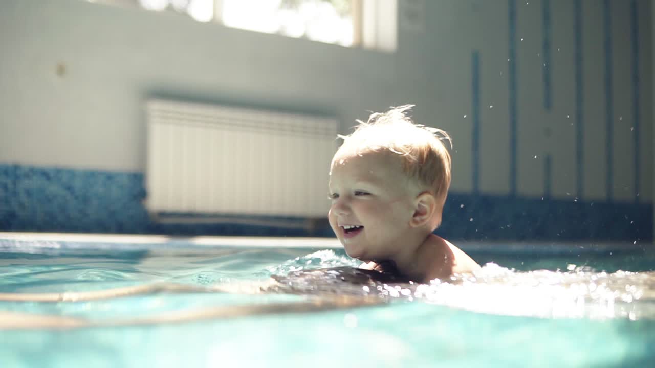 Young man dives under the water. Rowing in the closed swimming pool. Little boy is onhis fathers back. Smilling. Mother is hugging them. Happy family time