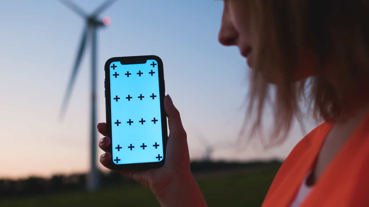 mujer ingeniera sosteniendo un teléfono inteligente con llave chroma y haciendo zoom en gesto en el fondo de la turbina eólica al atardecer.