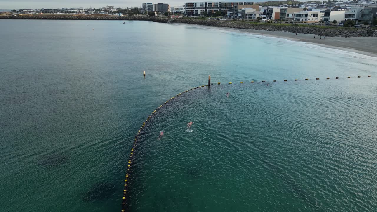 보호 구역 내에서 해가 뜨는 동안 바다에서 스포츠 운동을 하는 수영 선수의 드론  ⁇ 도 촬영 - 배경에는 퍼스 시의 럭셔리 빌라와 해변이 있습니다.
