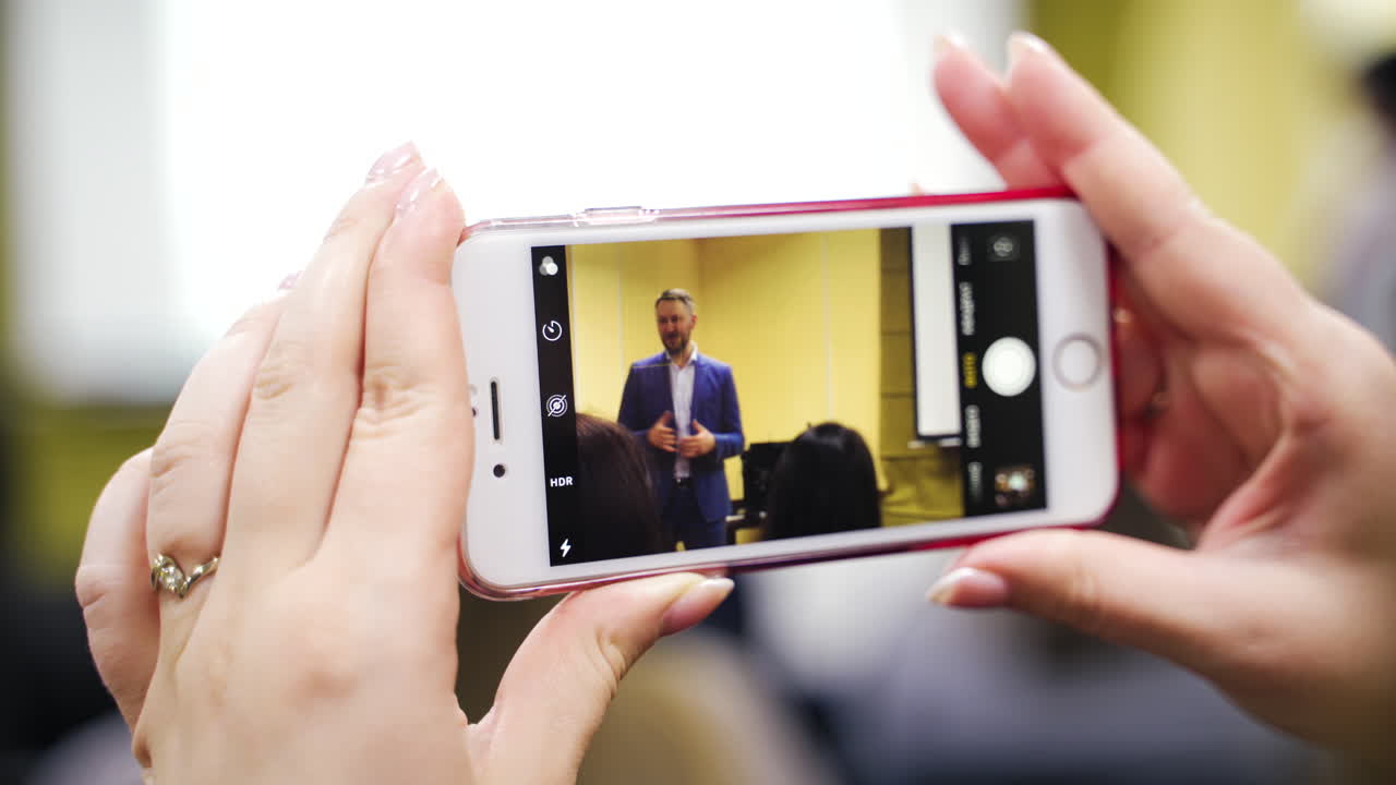 Smartphone in woman's hands during seminar. Woman is recording the video of the male speaker on her phone at conference indoors. Close-up.