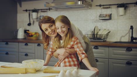 portrait shot of the beautiful mother hugging her pretty little daughter and they both smiling the camera in the kitchen at the table while baking. inside