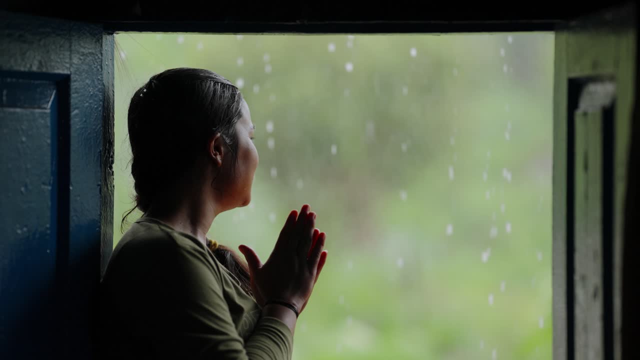 Happy Indian girl sitting by the door enjoying rainfall, peaceful rural life moment during monsoon, 4k video