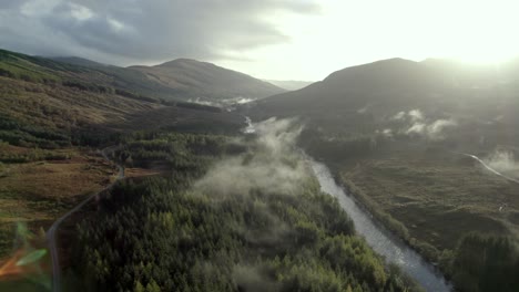 Luftaufnahmen-Von-Drohnen,-Die-Hoch-über-Einem-Fluss,-Einer-Straße-Und-Einem-Nadelwald-Fliegen,-Während-Tief-Hängende-Wolken-Die-Baumkronen-Umarmen-Und-Die-Sonne-Hinter-Bergen-Untergeht