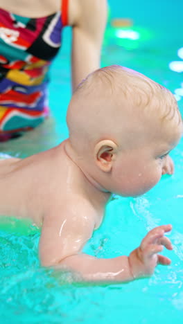beautiful caucasian baby swimming cheerfully in the swimming pool. unrecognized coach splashes water on child's back. vertical video.