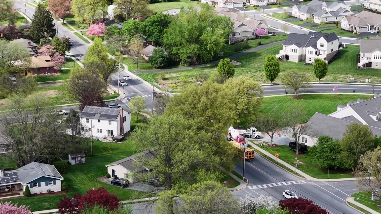 Aerial drone view above sunny American suburb in spring with yellow school bus moving through neighborhood roads. Houses with solar panels, green lawns and blooming trees. Peaceful residential scene.