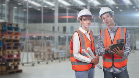 two caucasian workers in a warehouse with oranges high vest and hat area talking and checking notes