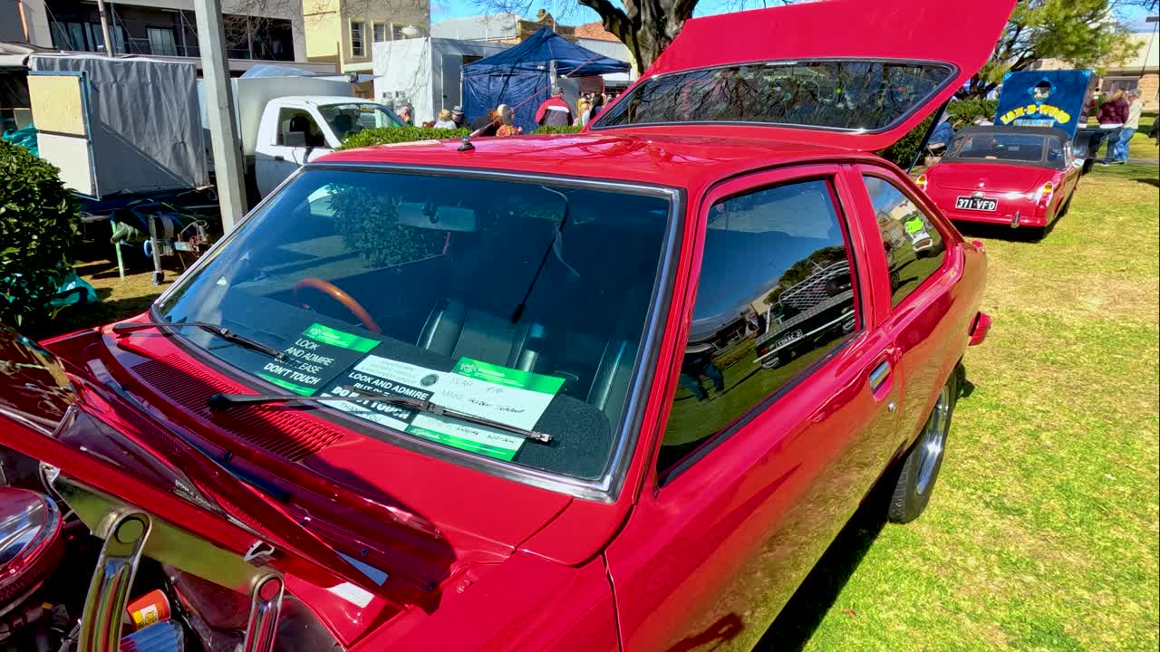 A classic red car’s engine bay is revealed in bright daylight, with camera panning outward to show the vehicle at an outdoor festival setting
