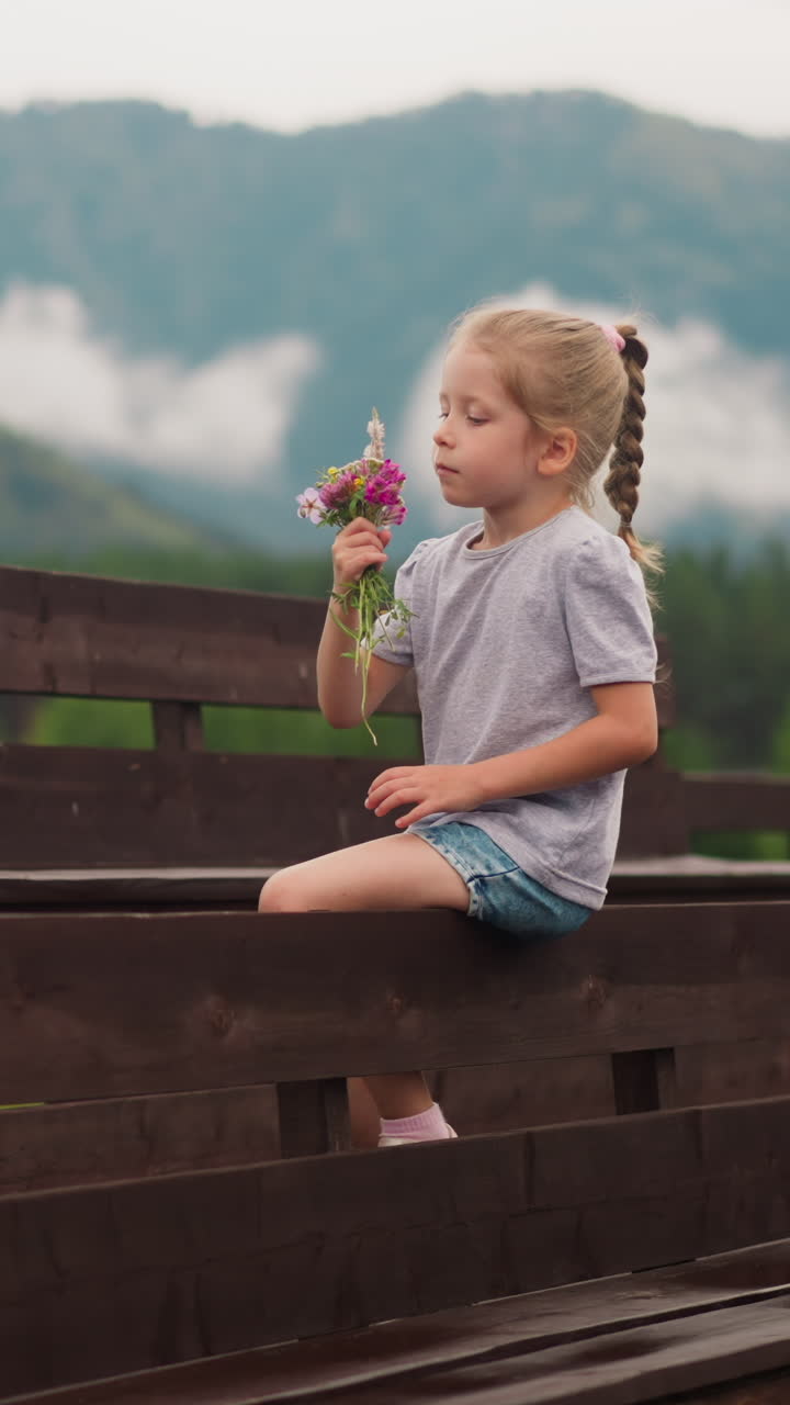 Cute little girl with plait enjoys aroma of wildflowers bouquet sitting on wooden bench against misty mountain silhouettes on spring day