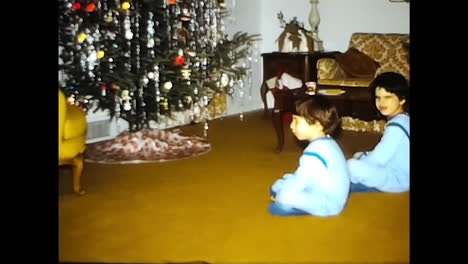kids sitting on floor in front of christmas tree. circa usa - 1970s: two children from a 1970s video archive in the usa are sitting on the floor in front of a christmas tree.