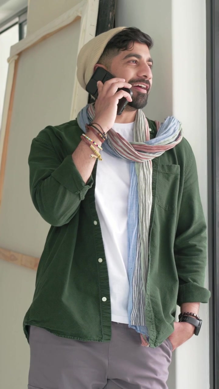 Vertical video: Taking call to chat, man leaning by door talking on phone in beanie, canvas nearby