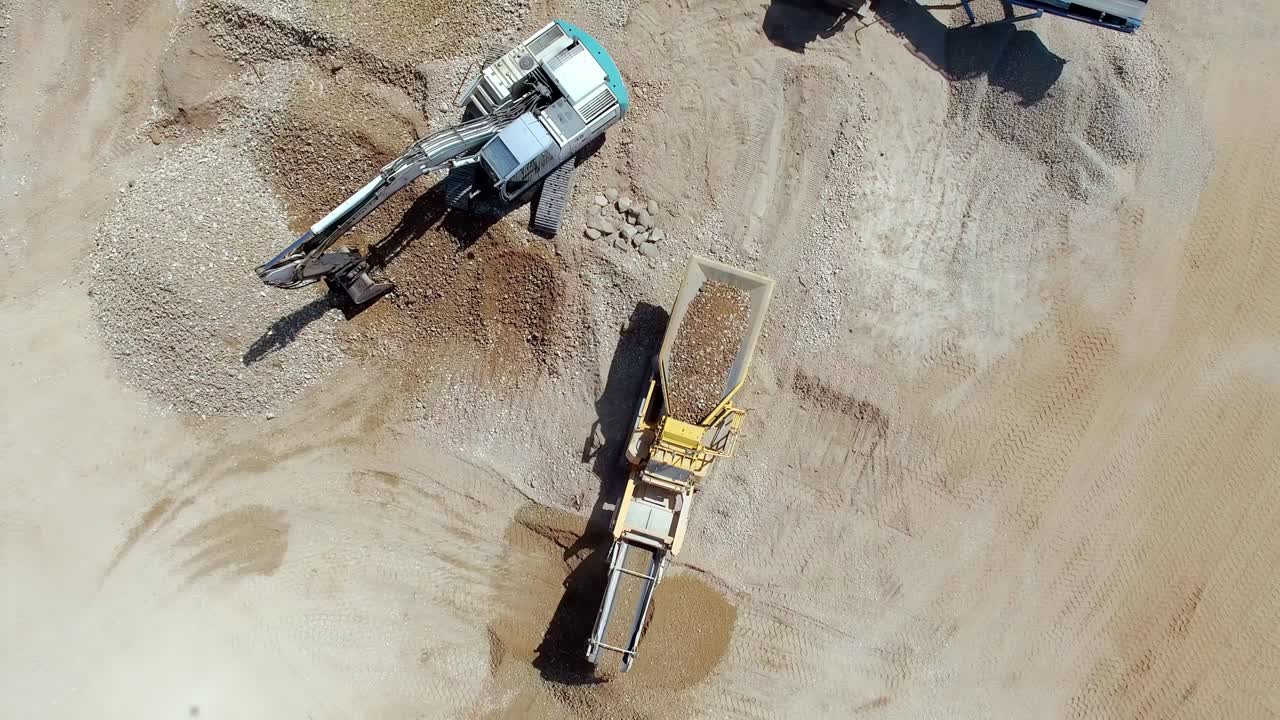 Excavator loading gravel into large industrial crusher at open pit mining site aerial view