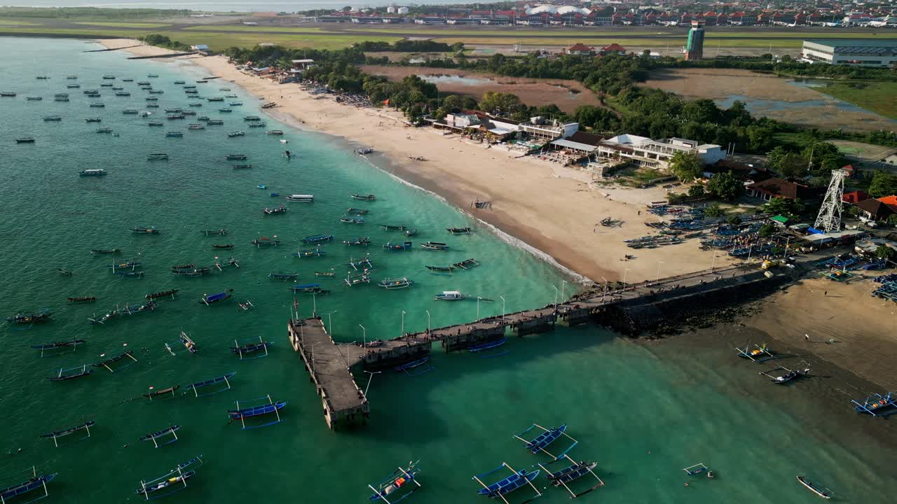 Aerial view of Bali pier surrounded by fishing boats floating in turquoise water, coastal shoreline stretching wide, and the harbor alive with daily island activity