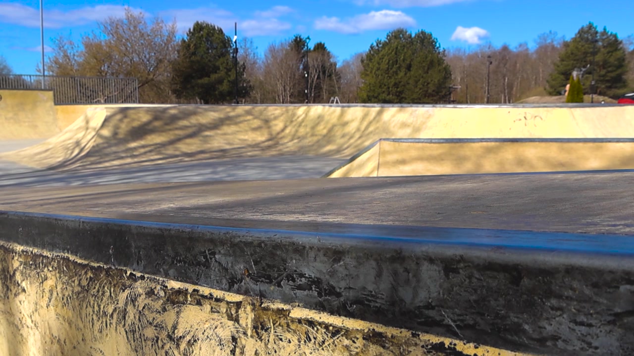 Movimento fluido della telecamera lungo una sporgenza di cemento in uno skatepark vuoto in una giornata di sole splendente, con alberi e un cielo blu sullo sfondo. Scena ideale per concetti di sport estremi e cultura giovanile.
