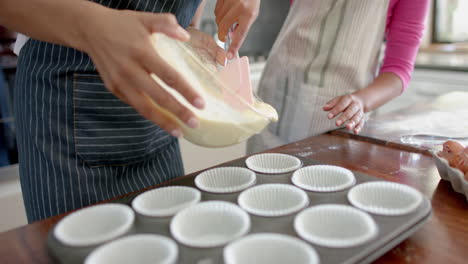 Midsection-of-biracial-mother-and-daughter-pouring-cake-mix-into-cake-forms-in-kitchen,-slow-motion