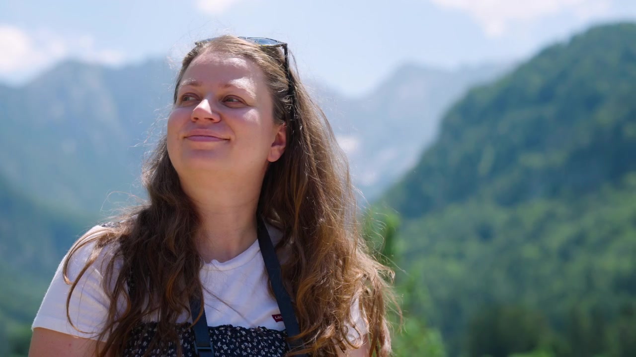 Smiling Woman Holds A Camera To Her Eye, Taking A Photo Against A Scenic, Blurred Green Mountain Backdrop. - medium shot