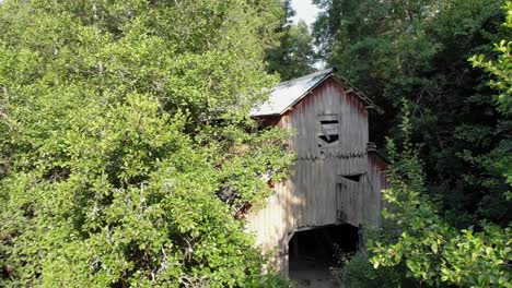 Backward-aerial-of-large-wooden-barn-by-lush-green-forest-in-sunlight