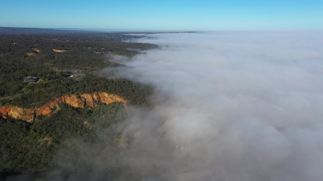 Una ripresa aerea con drone cattura un paesaggio in cui la nebbia avvolge una foresta e delle scogliere, con un cielo blu visibile sopra. Il drone effettua una panoramica sulla scena, rivelando la bellezza naturale dell'ambiente.