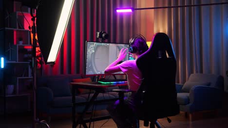 vista trasera de una streamer asiática que se estira mientras juega un juego a través de la red en una computadora personal. videojuego de transmisión en vivo, escritorio iluminado por luz de tira de led rgb