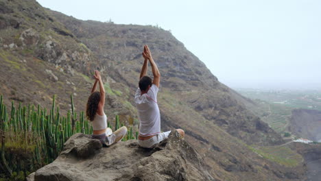 High-on-a-mountain,-a-man-and-a-woman-sit-on-stones,-meditating-with-raised-hands,-absorbing-the-ocean-view,-and-practicing-calming-breaths