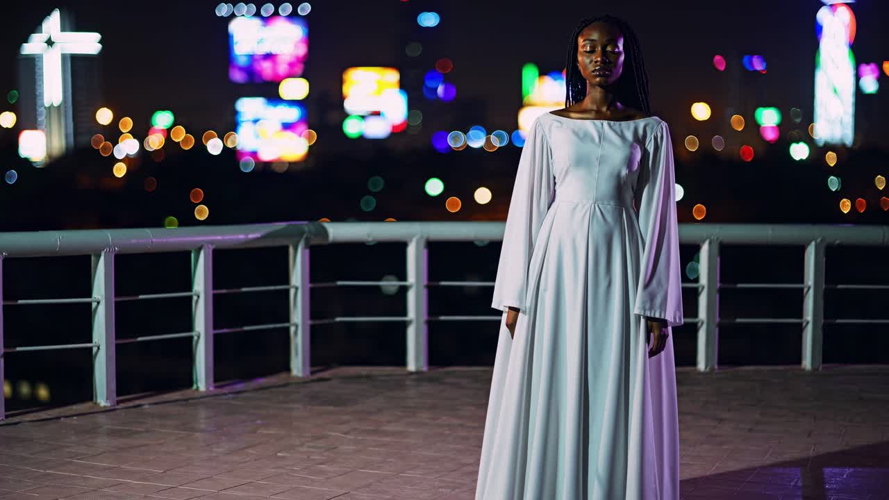Elegant woman in flowing white dress stands confidently on rooftop at night, illuminated by vibrant city lights, showcasing grace and poise in a dynamic urban setting