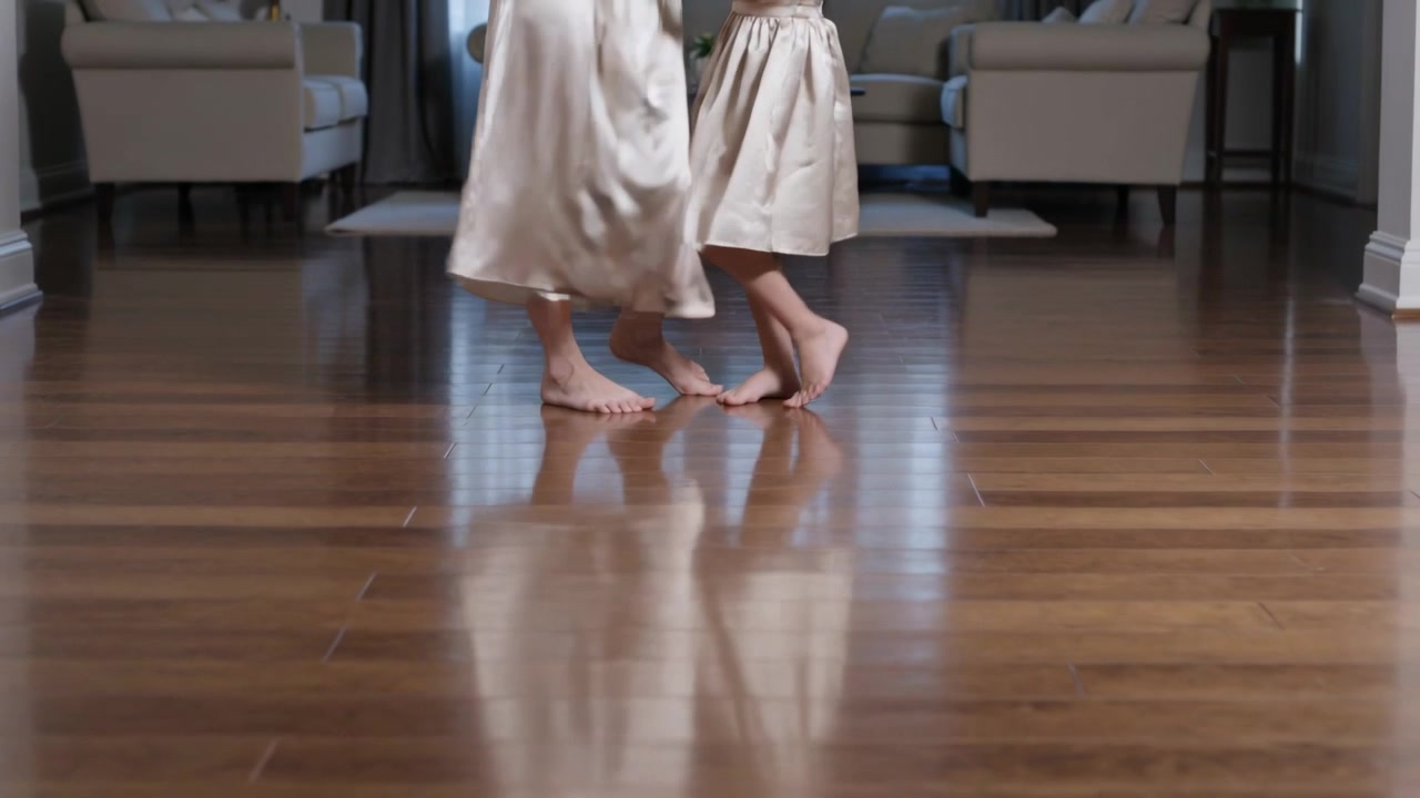 Shifting weight mom leading girl at home, practicing steps to bond on shiny floor in dresses