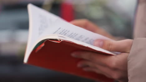 Close-up view of lady's hands holding and flipping pages of red book with blurred background, capturing quiet moment of reading outdoors in autumn setting with soft coat sleeves partially visible