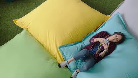 happy woman student using smartphone browsing online social media messages enjoying lunch break relaxing on colorful pillows listening to music in modern office top view