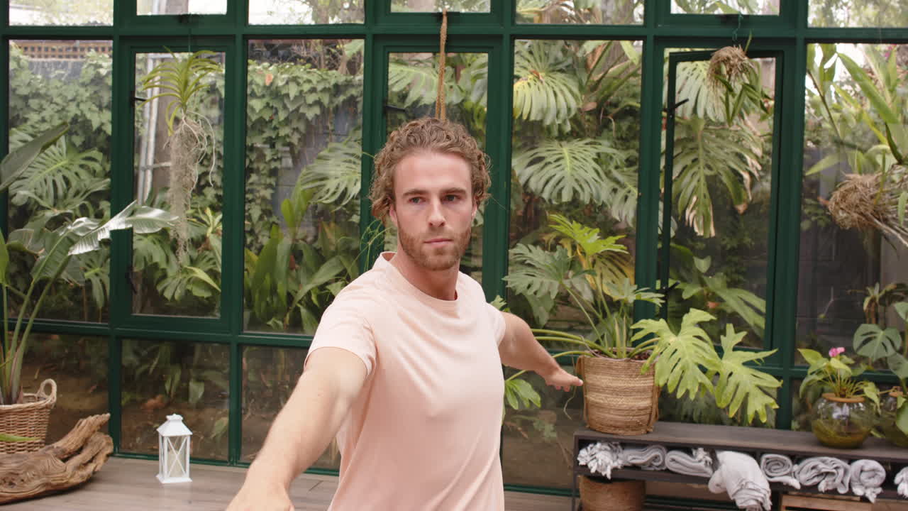 Practicing yoga, man stretching arms in warrior pose in glass house