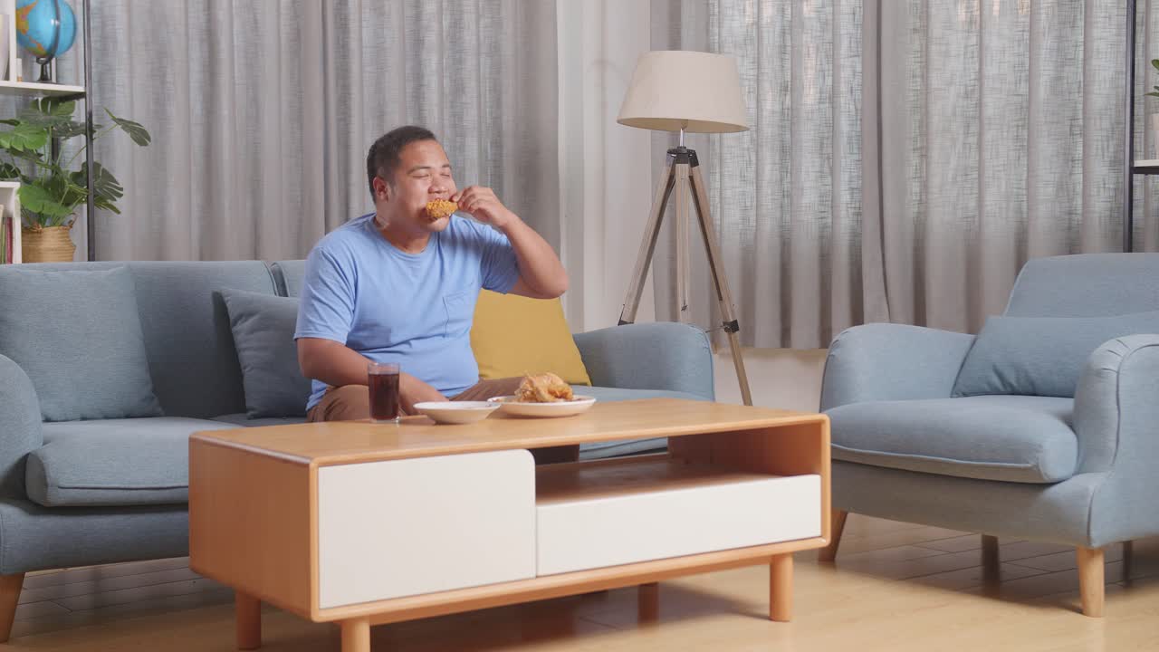 한 아시아 남자의 몸이 가득 차 있습니다. 집에서 거실에서 소파에 김 을 먹고 패스트푸드를 먹는 동안 코카콜라 한 잔을 마시고 있습니다.