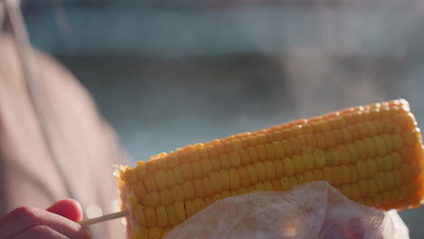 close up of person rotating corn while steam rises from freshly cooked corn, outdoors, blurred background showing chilly winter environment, healthy hot snack, warmth in cold weather