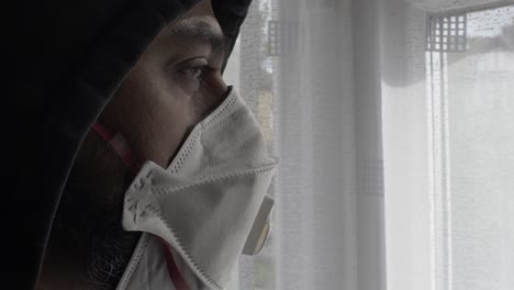 hooded adult asian male wearing ffp three mask indoors looking through curtain