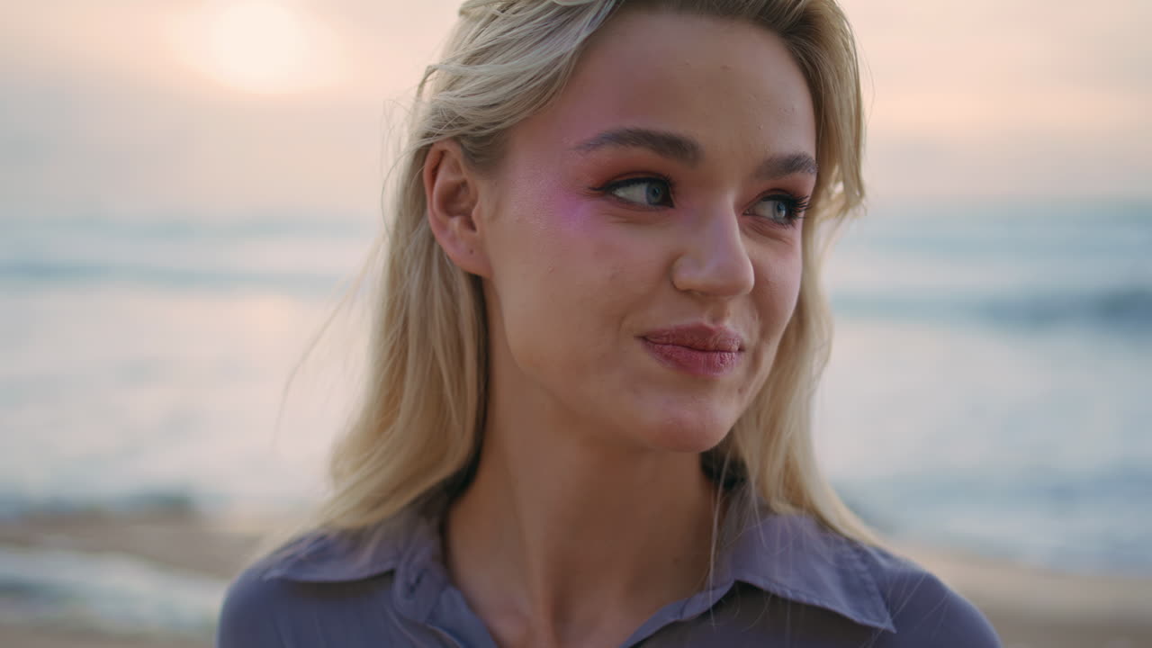 Smilende kvinde poserer ved havets solnedgang tæt på. Afslappet smuk blondine justerer håret