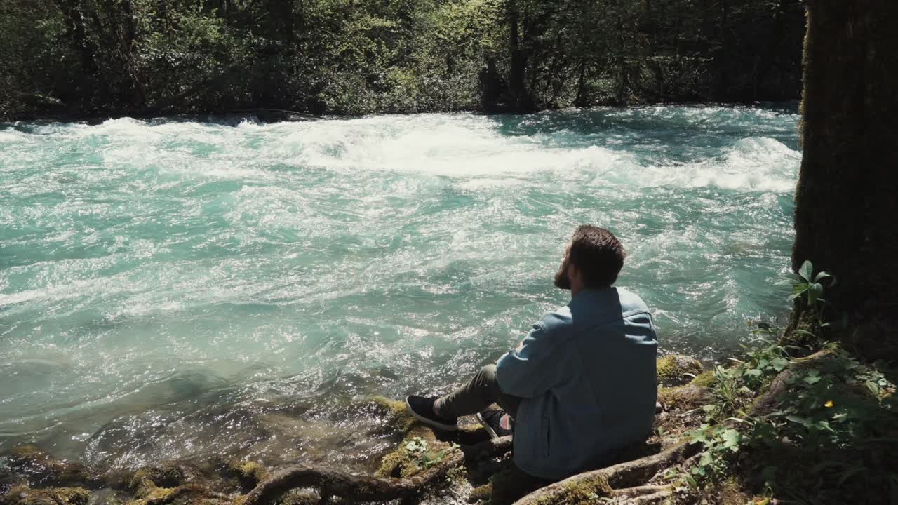 hombre sentado junto a un río turquesa en un bosque