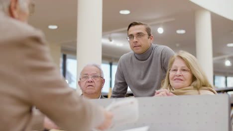 Senior-man-giving-textbook-to-woman-during-class-in-library