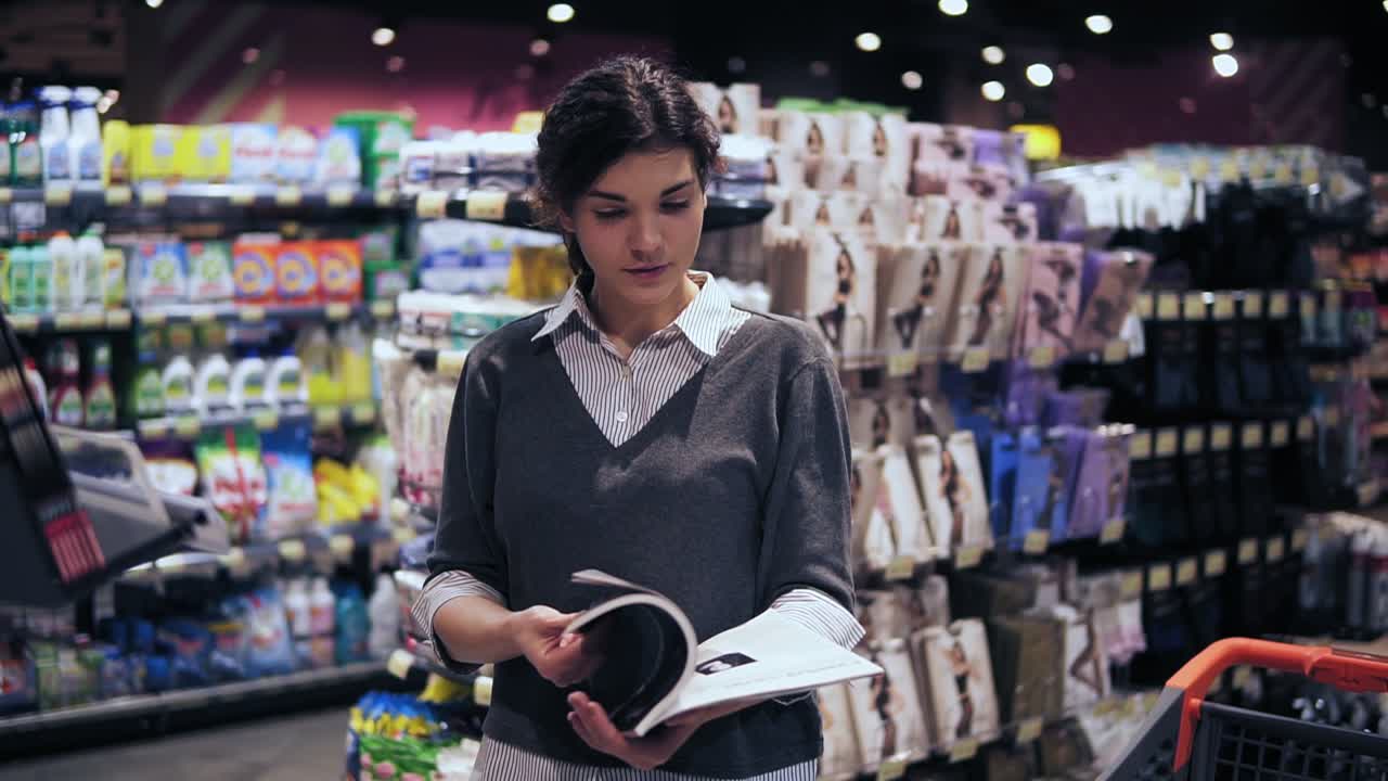 Young beautiful brunette girl at her 20's leafing through magazine in local supermarket