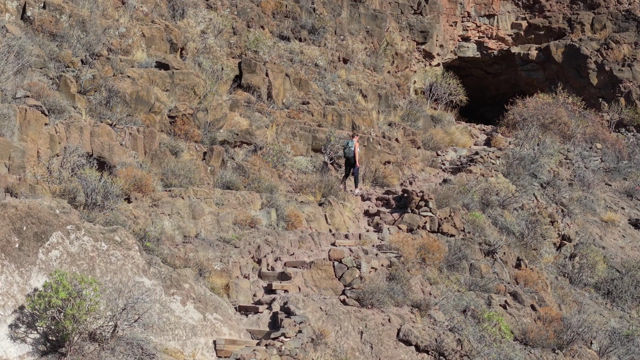Alone, a woman hiker makes her way down a narrow stone trail etched into the side of a rugged mountain, Ansite fortress. The sun shines brightly, creating the perfect setting for an outdoor exploratio