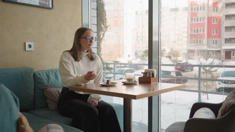 young woman in white sweater enjoys lunch by window in cozy cafe, holding spoon near dessert while seated on teal sofa, snow covered cars and buildings visible outside through glass panels
