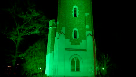 Beaumont-Tower-on-the-campus-of-Michigan-State-University-lit-up-at-night-in-green-in-honor-of-the-victims-of-the-February,-2023-mass-shooting-with-close-up-video-tilting-to-clock