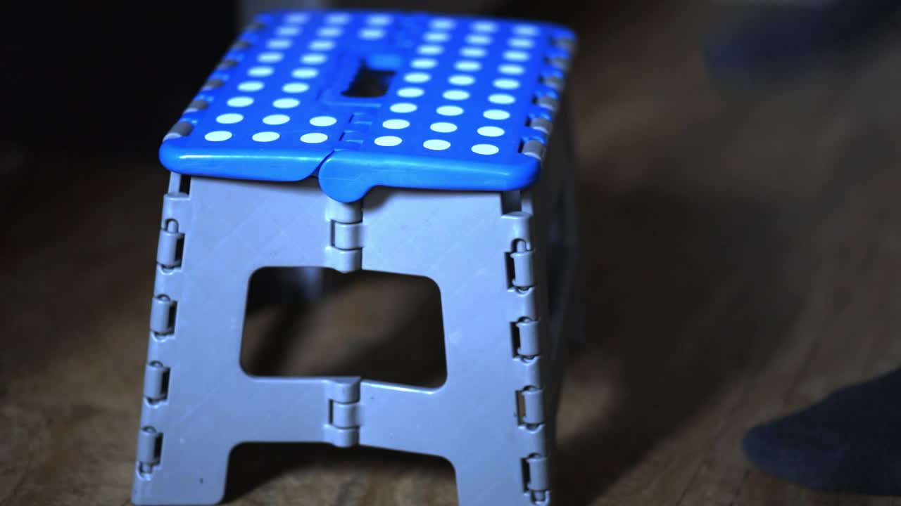 Person Stepping On A Blue Plastic Stool To Reach For Something On A Cabinet Counter In The Kitchen. - close up shot