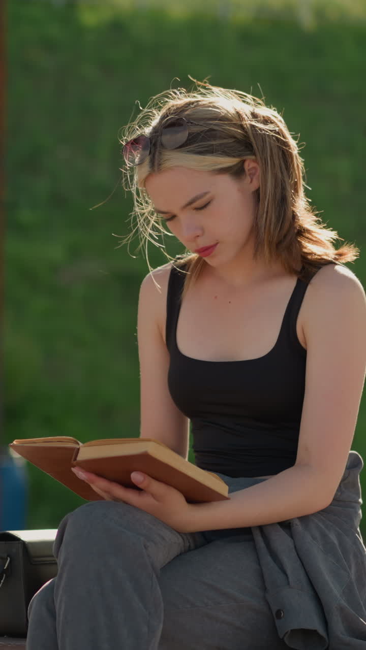 dama sentada con las piernas cruzadas leyendo un libro. el viento sopla su cabello mientras su mano descansa en el libro, una bolsa negra está a su lado, con un fondo borroso con vegetación y estructuras de madera