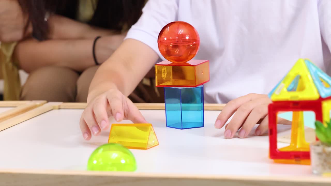 Child builds tower with translucent blocks at table, bright lighting, teacher and classmates observing